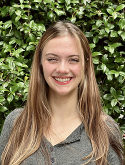 Smiling young woman with long hair standing in front of a green leafy background.