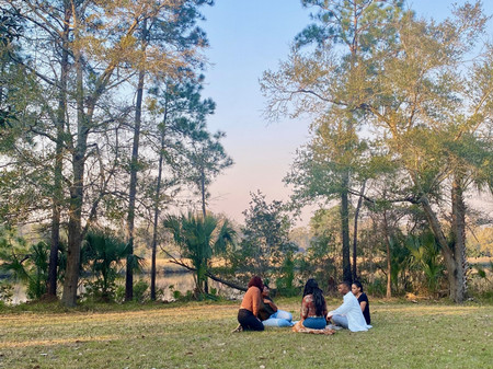 Group of five students sitting in open field in front of trees and a pond. 