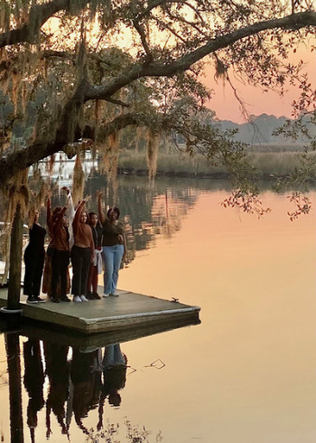 Six students standing on a dock at sunset with their hands in the air.