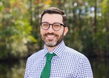 A smiling man with glasses and a green tie stands in front of a blurred natural background.