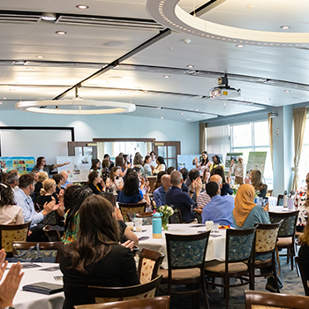 Large room of round tables with individuals sitting.