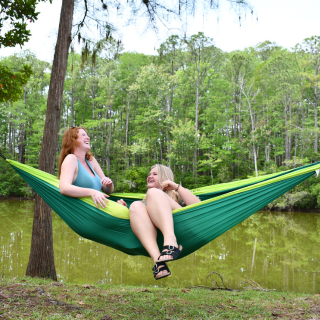 Two people relaxing in a green hammock by a forested lake.
