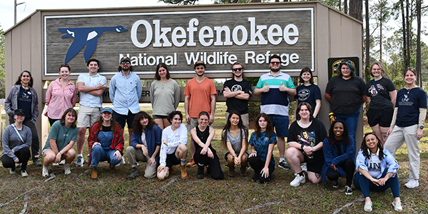 Twenty three individuals posing for a photo in front of a sign.
