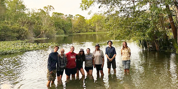 Eight individuals standing knee high in lake posing for photo.
