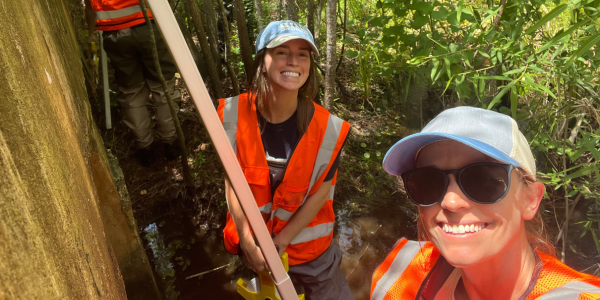 Two field researchers in safety vests surveying a wooded creek