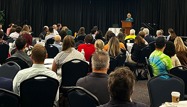 Auditorium with a speaker on stage presenting to attendees sitting at round tables.