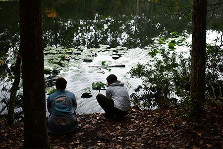 Two people sit at the edge of a calm lake.