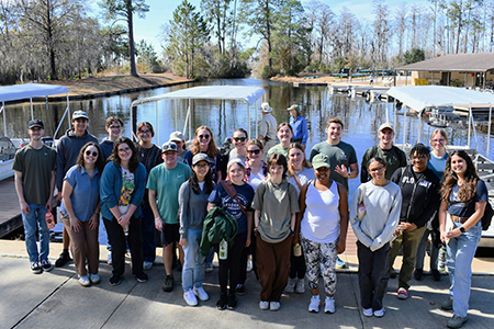 Team poses by dock on sunny day with boats and trees behind.