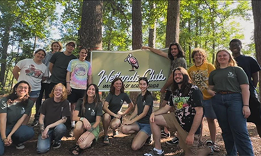 Group of students standing in a forest preserve with a banner featuring the wetlands club.