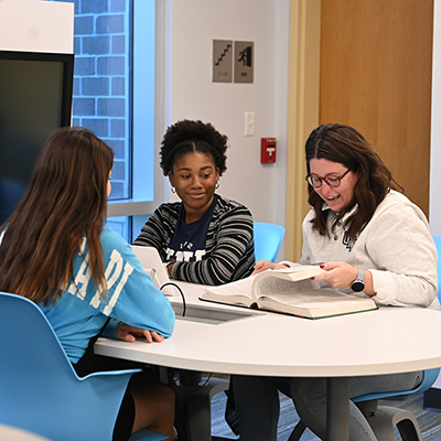 Three students in UNF Library sitting at a table looking over a book 