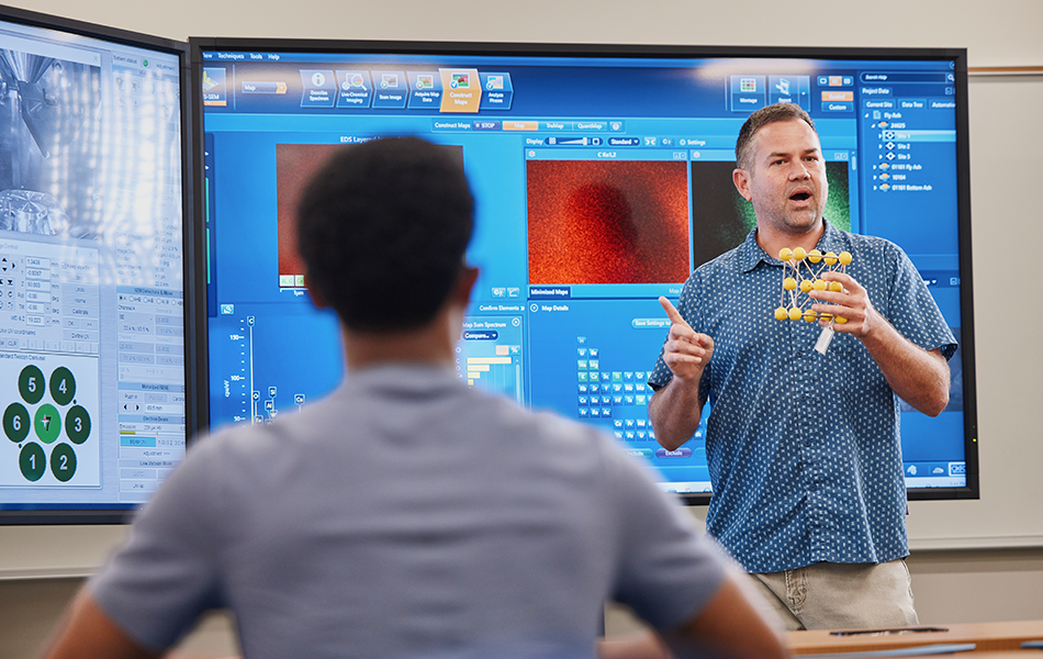 Professor in front of a monitor and holding a scientific model while giving lecture to a student