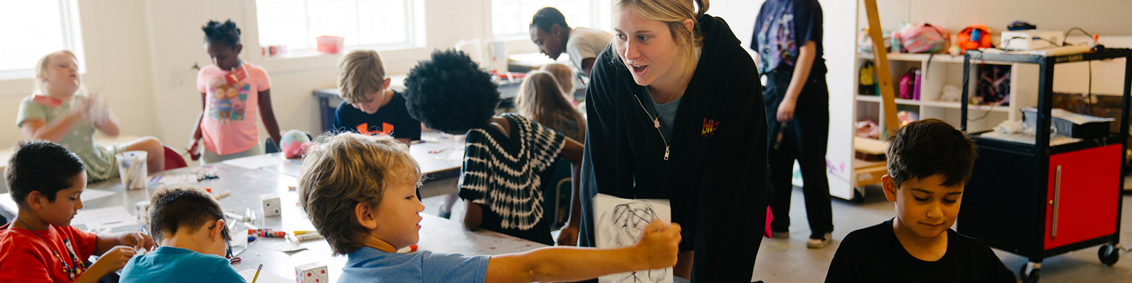 Children engaged in an art class, interacting with a teacher as they create projects at their desks.