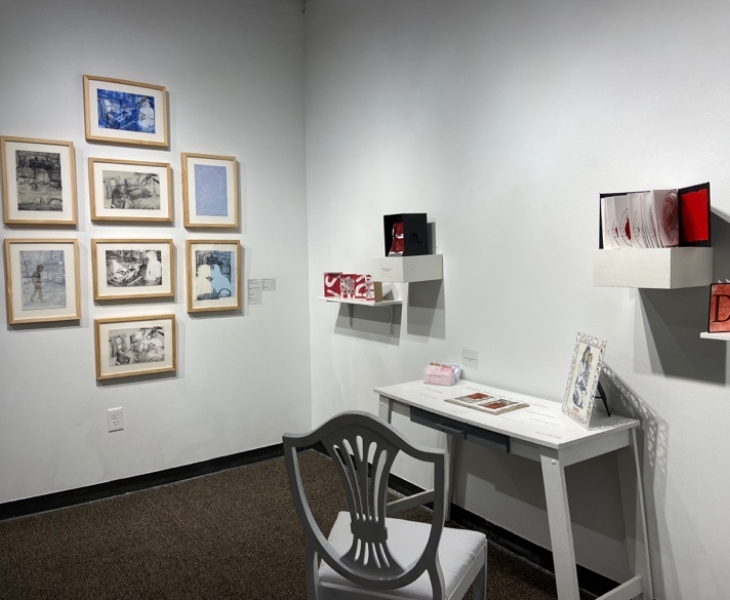 Gallery installation with framed artwork, wall-mounted pieces, and a white desk and chair.