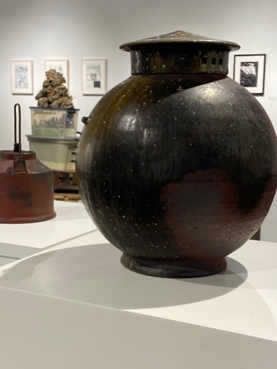 Dark spherical ceramic vessel displayed on a white pedestal in a gallery.