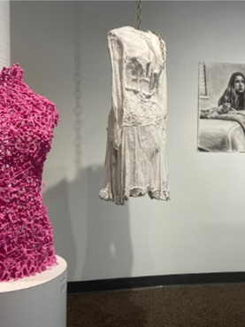 Pink textured torso sculpture, hanging white garment, and black-and-white artwork displayed in a gallery.