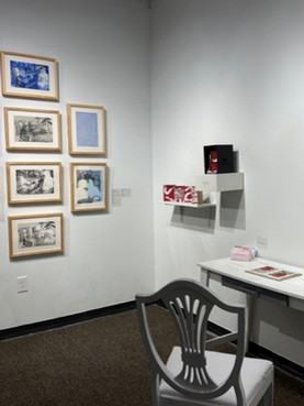 Gallery installation with framed artwork, wall-mounted pieces, and a white table and chair.