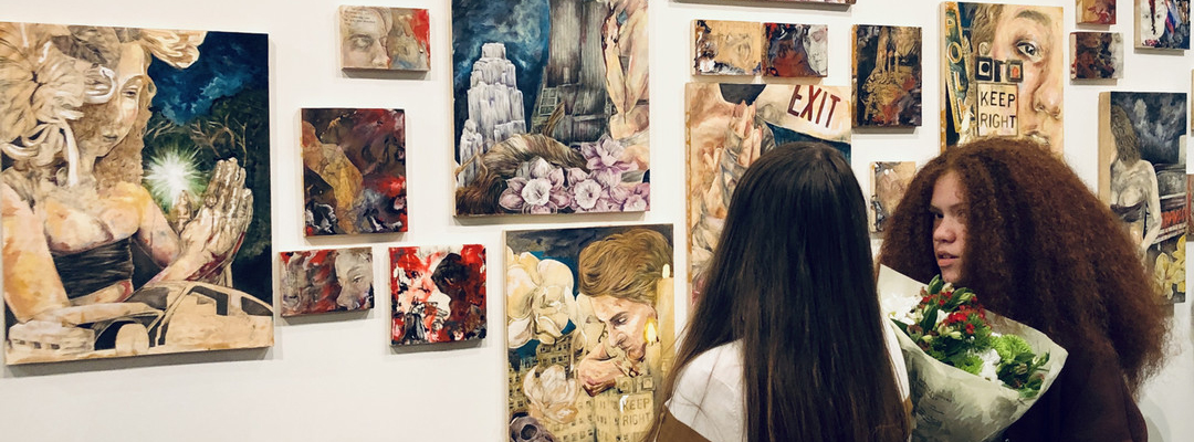 Two women admire a collection of vibrant mixed media artworks displayed on a gallery wall.