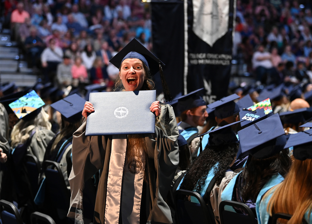 graduating student holding diploma folder and standing in crowd of sitting graduates