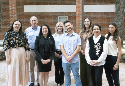 A group of graduate students, a dean, associate dean, director, and assistant director smile together outdoors in front of a brick building on campus.