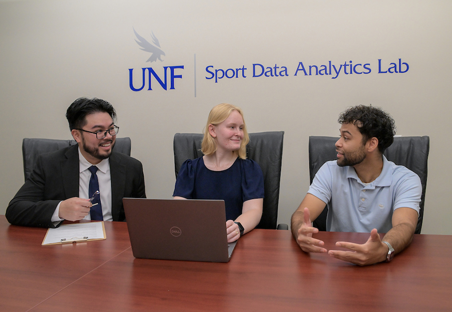 Three UNF students sitting in front of a UNF Sport Data Analytics Lab sign