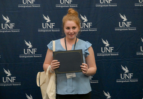 Blond woman in blue blouse holding award at UNF Student Research Symposium