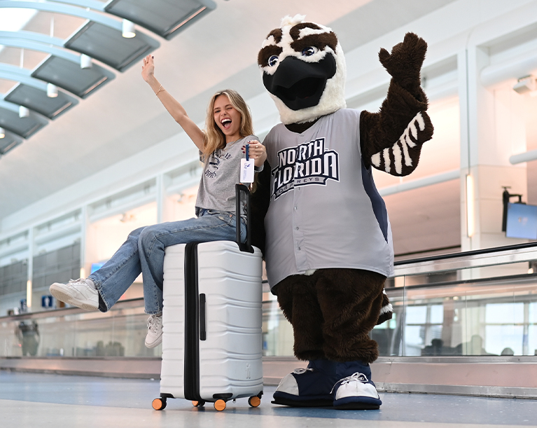 UNF student sitting on a suitcase cheering with the UNF mascot in the airport