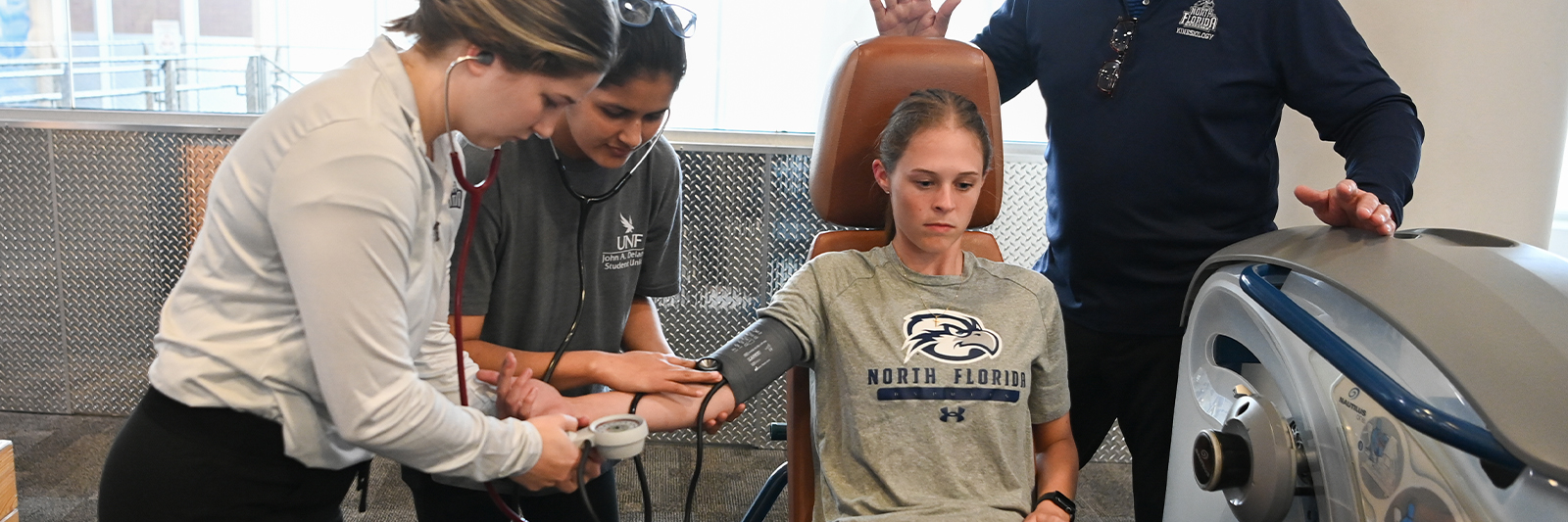 Two UNF students checking blood pressure while patient exercises in a gym