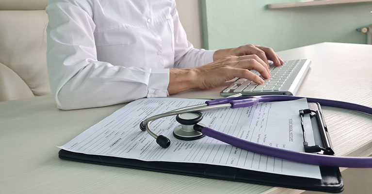 woman typing on a keyboard with a form on a clipboard and stethoscope