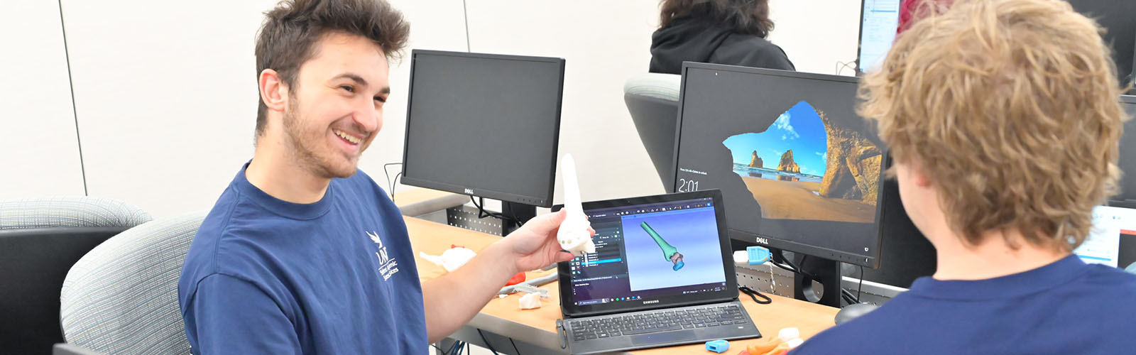 a student holding a 3-D printed bone with a computer model on a laptop talking to another student