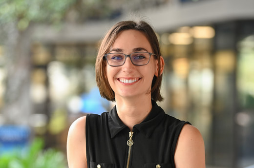 White female student with black shirt and blue glasses smiles in front of blurry background. 