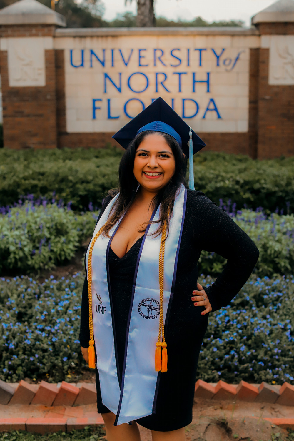 Girl in black dress and wearing a dark blue graduation cap stands in front of a UNF sign.