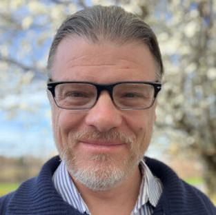 Headshot photo of a main with glasses outside. 