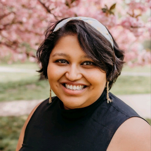 Nishi Dsouza smiling in front of a pink floral background.