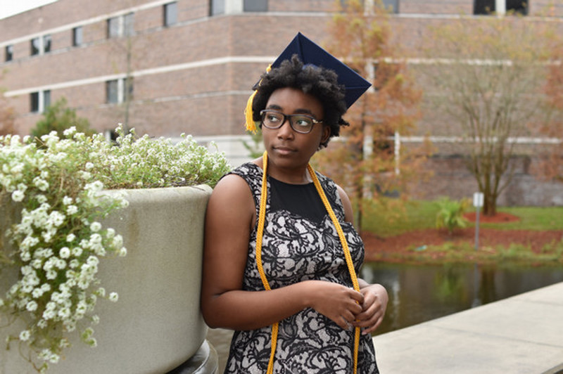 black female student poses in front of lake with blue graduate cap and yellow honor chords.