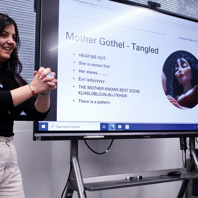 A student smiles while standing in front of a power point slide featuring Mother Gothel from Disney's Tangled.