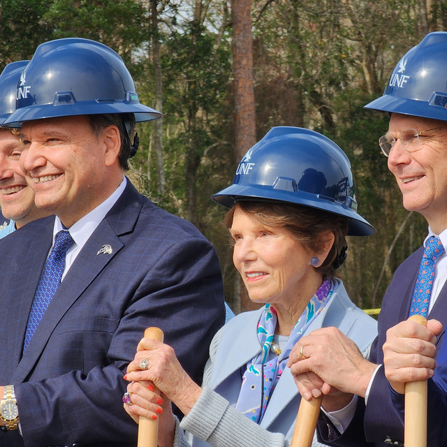 Mrs. Hicks and President Limayem smiling and wearing dark blue hardhats with woods in the background.