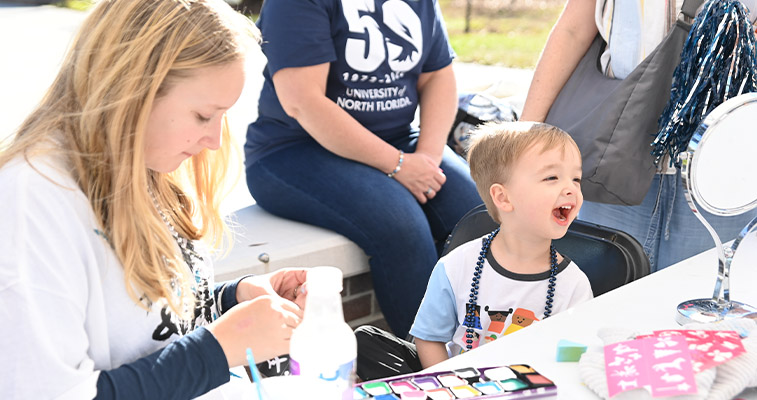 A child smiling while waiting to get his face painted by a volunteer