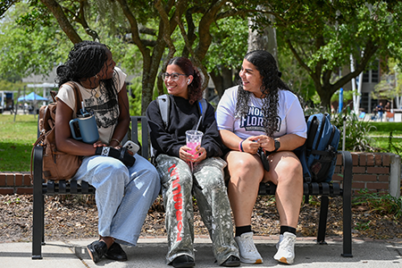 three UNF student sitting on a bench outside on UNF campus talking on a beautiful day