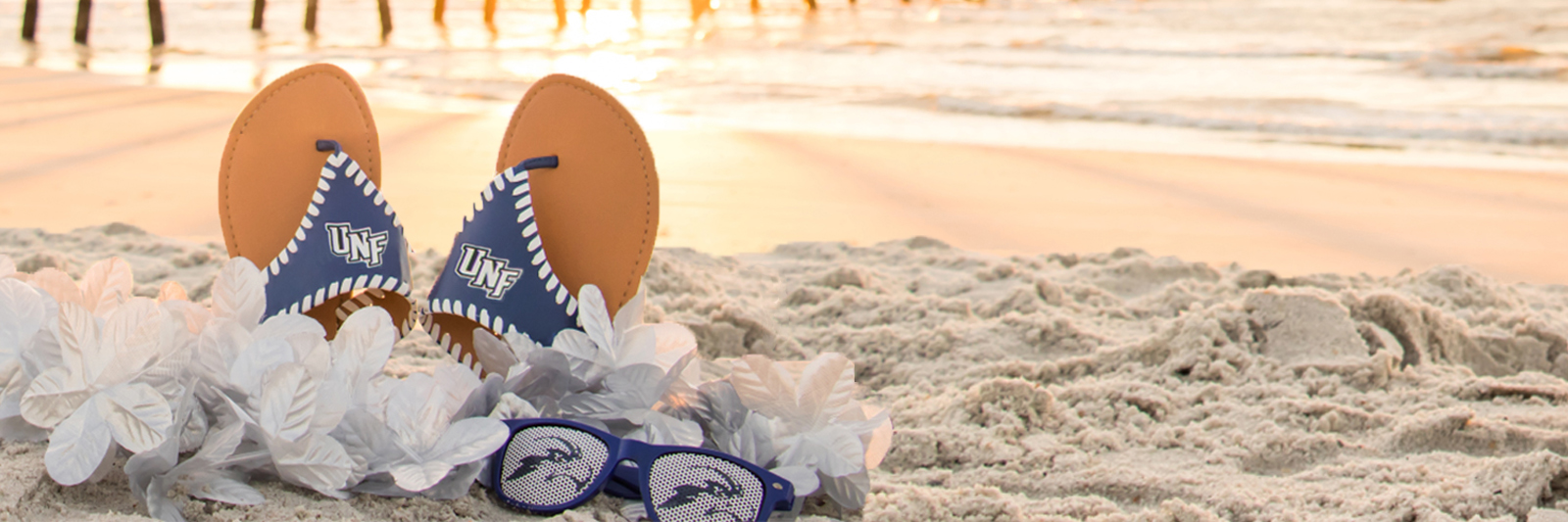 File: Pair of UNF flip flops, lei necklaces, and sunglasses on the beach with pier in background