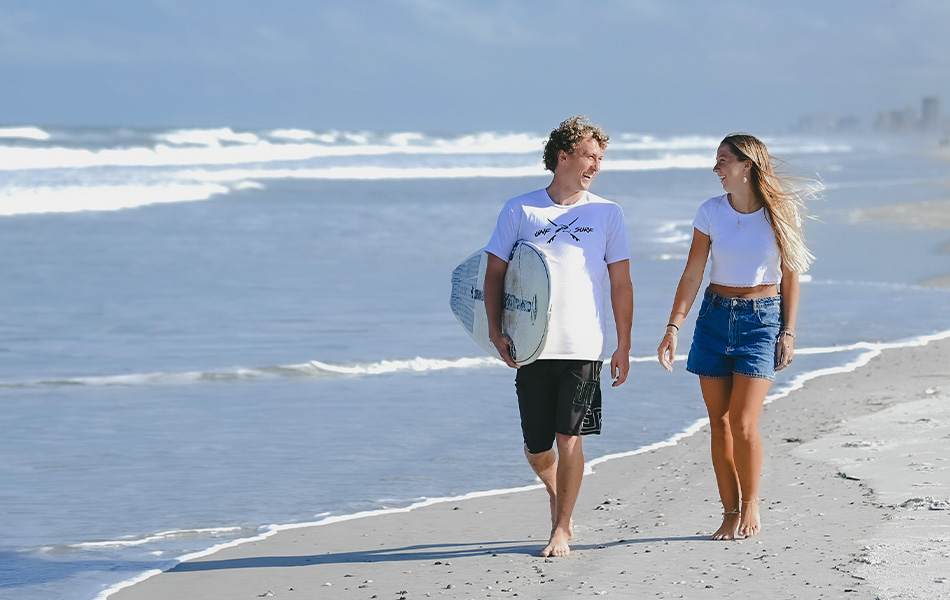 Two students walking on the beach while one holds a surfboard