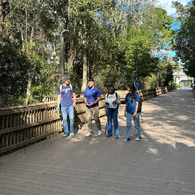 four students walking on the boardwalk with trees surrounding them