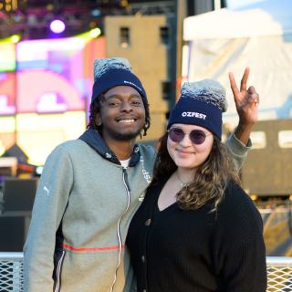 two students stand in front of a music stage and smile. One student holding up a peace sign.