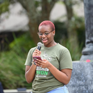 Student is holding a microphone and speaking into it while also holding a cell phone. 