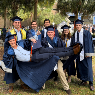 7 students smile wearing graduation caps and gowns. 6 of the students are standing and holding one student. 