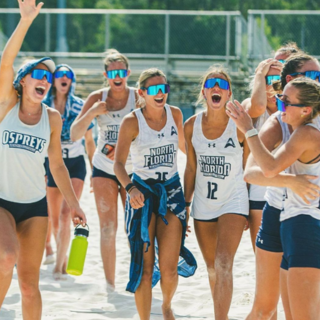 Volleyball players wearing uniform and sunglasses cheer and smile while standing in sand. 