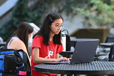 Female UNF student in a red shirt sitting outside looking at a laptop