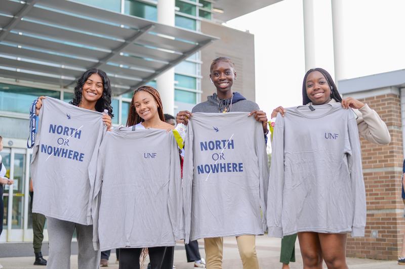 Four female UNF students holding up North or Nowhere shirts in front of the UNF library