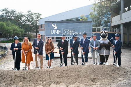 UNF president and employees wearing hard hats and holding shovels in front of a Coggin College of Business sign
