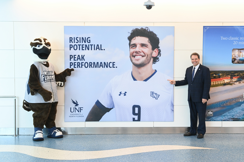 Ozzie and President Limayem stand in front of a UNF ad at the Jacksonville Airport