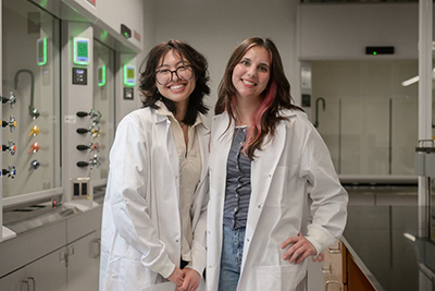 Two UNF students in white lab coats smiling inside of a chemistry lab